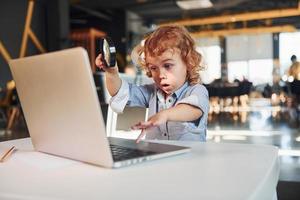 Smart child in casual clothes with laptop on table have fun with magnifying glass photo