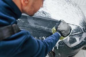 Male worker in uniform washing new modern car that is full of soap. Conception of service photo