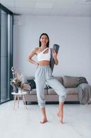 Standing and holds mat. Young woman with slim body shape in sportswear have fitness day indoors at home photo