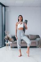 Standing and holds mat. Young woman with slim body shape in sportswear have fitness day indoors at home photo