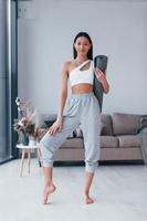 Standing and holds mat. Young woman with slim body shape in sportswear have fitness day indoors at home photo