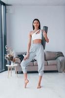 Standing and holds mat. Young woman with slim body shape in sportswear have fitness day indoors at home photo