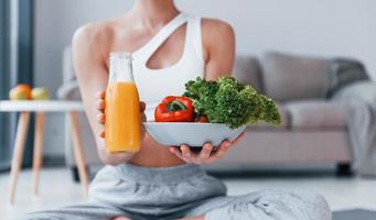 con comida saludable y jugo de naranja en las manos. mujer joven con forma de cuerpo delgado en ropa deportiva tiene un día de fitness en casa foto