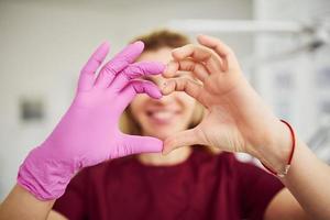 Young female dentist in uniform standing in stomatology office and making heart shaped gesture photo