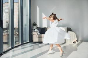 Cute little girl in white dress dancing indoors in bedroom alone photo