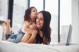 Young mother with her daughter spending weekend together in room and using laptop photo