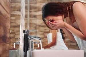 Beautiful young woman standing in bathroom near the mirror and washing her face. Friends behind photo