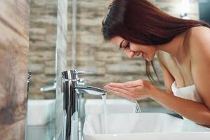 bella joven parada en el baño cerca del espejo y lavándose la cara foto