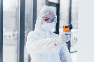 Holds infrared thermometer. Portrait of female doctor scientist in lab coat, defensive eyewear and mask photo