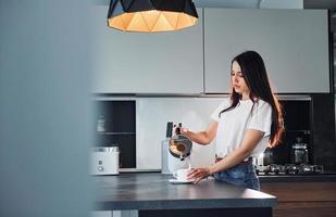 vertiendo leche fresca en el vaso. joven hermosa morena con ropa informal en el interior de la cocina durante el día foto