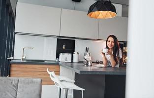 con taza de café recién hecho. joven hermosa morena con ropa informal en el interior de la cocina durante el día foto