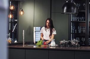 Vegetables and milk on table. Preparing food. Young beautiful brunette in casual clothes indoors in kitchen at daytime photo