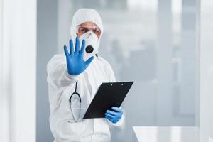 Shows stop gesture by hand. Male doctor scientist in lab coat, defensive eyewear and mask holding notepad in hands photo