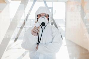 Female doctor scientist in lab coat, defensive eyewear and mask standing indoors and holds stethoscope photo