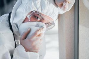 Feels bad and sick. Female doctor scientist in lab coat, defensive eyewear and mask standing indoors photo