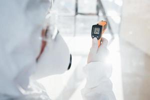 Female doctor scientist in lab coat, defensive eyewear and mask standing indoors with infrated thermometer photo