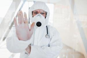 Stop the virus gesture. Female doctor scientist in lab coat, defensive eyewear and mask standing indoors photo