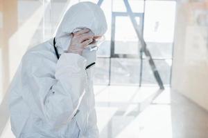 Feels bad and sick. Female doctor scientist in lab coat, defensive eyewear and mask standing indoors photo