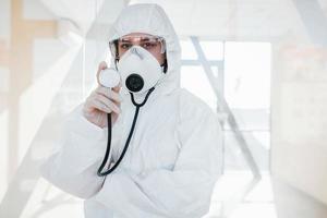 Female doctor scientist in lab coat, defensive eyewear and mask standing indoors and holds stethoscope photo