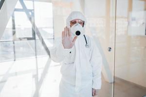 Stop the virus gesture. Female doctor scientist in lab coat, defensive eyewear and mask standing indoors photo