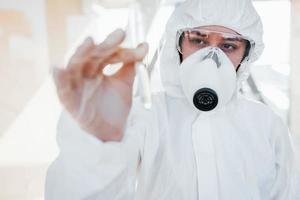 With test tube in hand. Female doctor scientist in lab coat, defensive eyewear and mask standing indoors photo