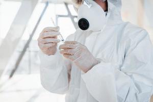 Holds syringe with medicine. Female doctor scientist in lab coat, defensive eyewear and mask standing indoors photo