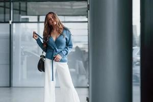 Attractive woman in blue shirt and white pants indoors posing for a camera. In airport or car shop photo