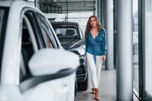 Positive woman in blue shirt walks near brand new car. In auto salon photo