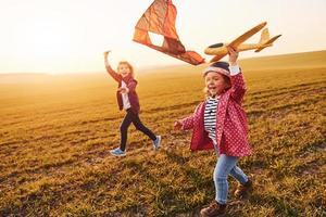 Two little girls friends have fun together with kite and toy plane on the field at sunny daytime photo