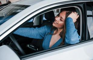 Positive woman in blue shirt sits inside of new brand new car. In auto salon or airport photo
