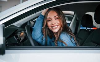 Positive woman in blue shirt sits inside of new brand new car. In auto salon or airport photo