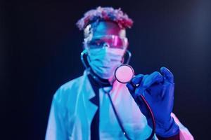 Smart doctor in protective uniform holding stethoscope. Futuristic neon lighting. Young african american man in the studio photo