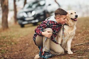 Cheerful boy in casual clothes sitting with her dog in forest against modern black car photo