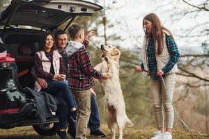 Happy family sitting and having fun with their dog near modern car outdoors in forest photo