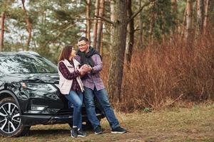 Positive mature couple standing near their car wit warm drink in hands. Outdoors in forest photo
