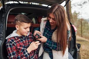 Sister pouring warm drink for her brother outdoors in forest when sits on back of car photo