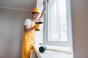 Handyman in yellow uniform installs new window by using automatic screwdriver. House renovation conception photo