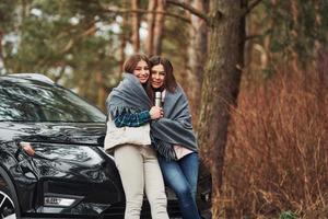 Mother and daughter standing together near modern black car outdoors in the forest photo