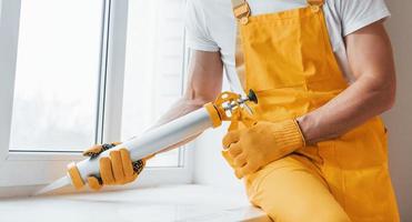 Handyman in yellow uniform works with glue for window indoors. House renovation conception photo
