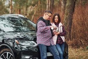 Positive mature couple standing near their car wit warm drink in hands. Outdoors in forest photo