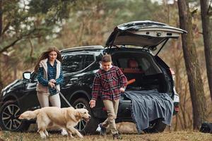 Cheerful girl with brother have walk with their dog outdoors in forest at autumn or spring season near car photo