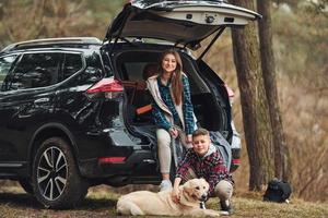 una chica alegre con su hermano se sienta en la parte trasera de un camión moderno con su perro al aire libre en el bosque foto