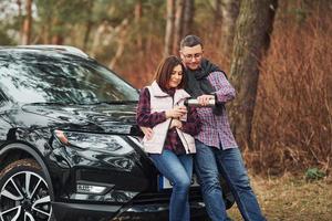 Positive mature couple standing near their car wit warm drink in hands. Outdoors in forest photo