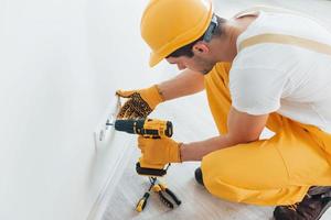 Handyman in yellow uniform works with electricity and installing new socket by using automatic screwdriver. House renovation conception photo