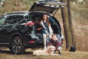 Cheerful girl with her brother sits on the back of modern car with their dog outdoors in forest photo