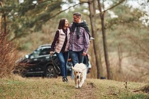 Happy mature couple have a walk with their dog in autumn or spring forest near modern car photo