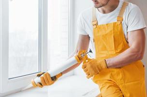 Handyman in yellow uniform works with glue for window indoors. House renovation conception photo