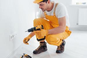 Handyman in yellow uniform works with electricity and installing new socket by using automatic screwdriver. House renovation conception photo