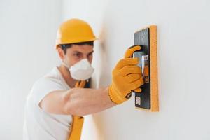 Handyman in yellow uniform and protective mask polishing wall indoors. House renovation conception photo