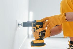 Handyman in yellow uniform works with electricity and installing new socket by using automatic screwdriver. House renovation conception photo